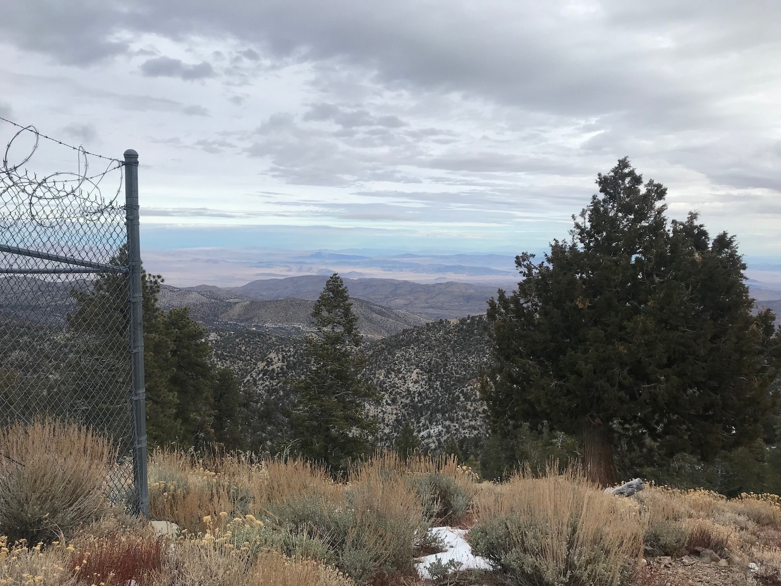 Scenic mountain overlook with layered hills, trees, and distant desert landscape under cloudy sky.