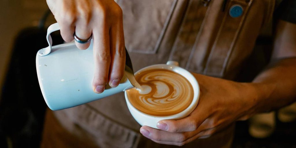 A person in an apron pours steamed milk from a metal pitcher into a white coffee cup, creating latte art at Big Bear.