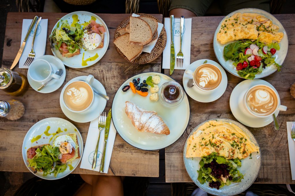 An overhead view of a wooden table at a restaurant in Big Bear, covered with various breakfast and brunch dishes, including omelets, avocado toast with smoked salmon, croissants, and multiple cups of lattes.