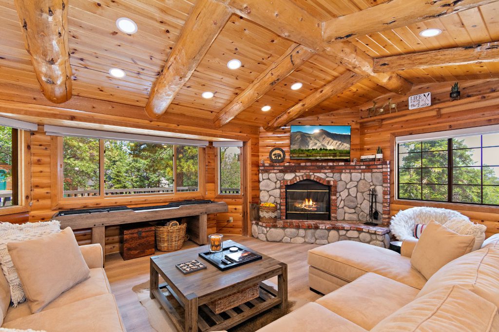 A cozy, rustic cabin living room in Big Bear with log walls and ceiling, a stone and brick fireplace, and a light-colored sectional sofa.