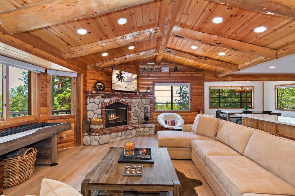 A cozy, rustic cabin living room in Big Bear with a large stone and brick fireplace, a light-colored sectional sofa, and a piano on the left.