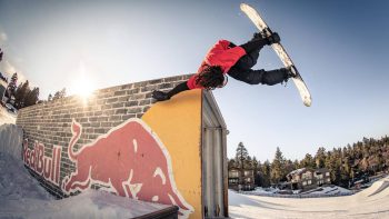 Snowboarder doing a wall ride trick on a Red Bull feature at Big Bear Mountain Resort, with snow and trees in the background.