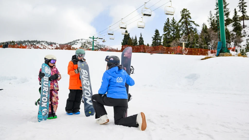 Snowboarding instructor teaching two kids on a snowy slope at Big Bear Mountain with chair lifts in the background under a cloudy sky