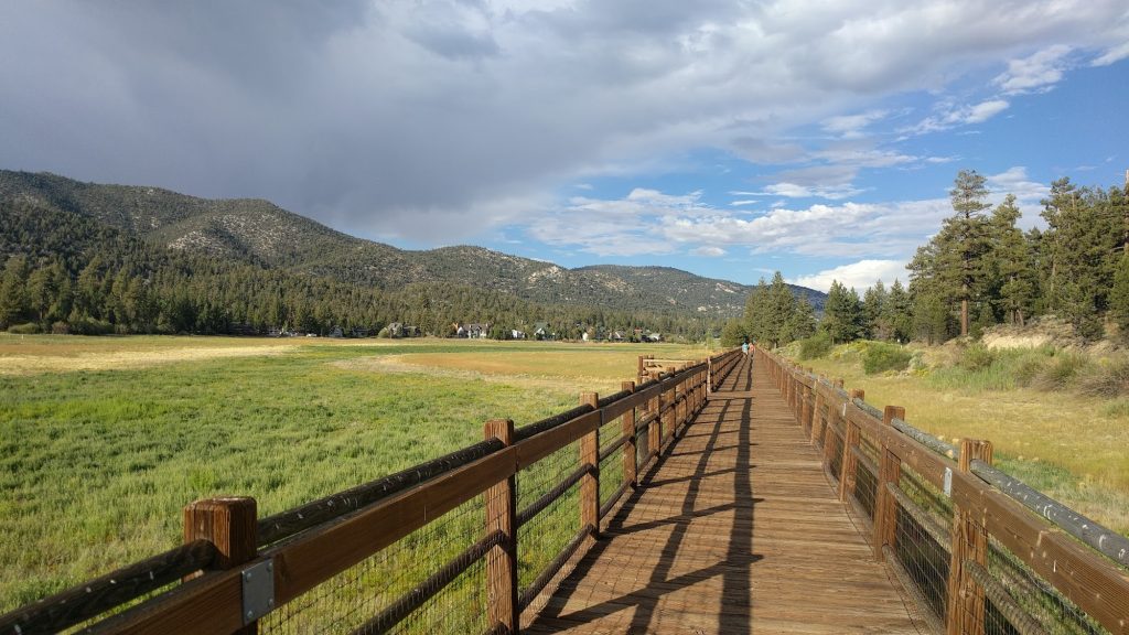 Wooden boardwalk stretching through the Stanfield Marsh wetlands in Big Bear Lake with mountain views, pine trees, and dramatic skies.