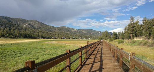 Wooden boardwalk stretching through the Stanfield Marsh wetlands in Big Bear Lake with mountain views, pine trees, and dramatic skies.