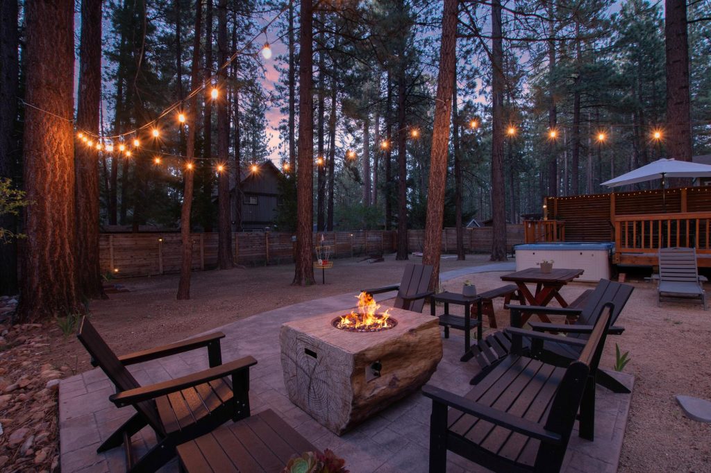 Backyard forest retreat in Big Bear featuring Adirondack chairs around a fire pit, string lights, tall pine trees, and an outdoor hot tub at dusk.