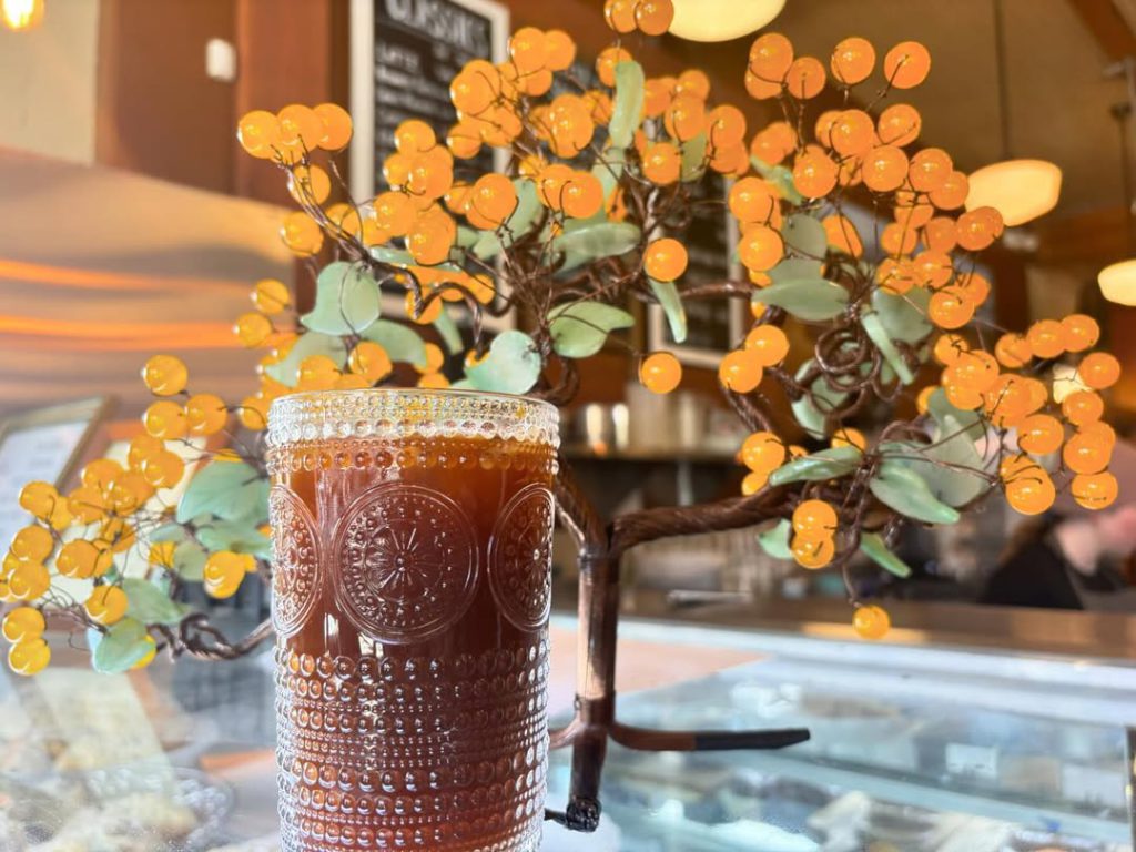 A glass of iced coffee with a decorative pattern, placed in front of a tree-shaped sculpture with orange glass balls at a restaurant in Big Bear.