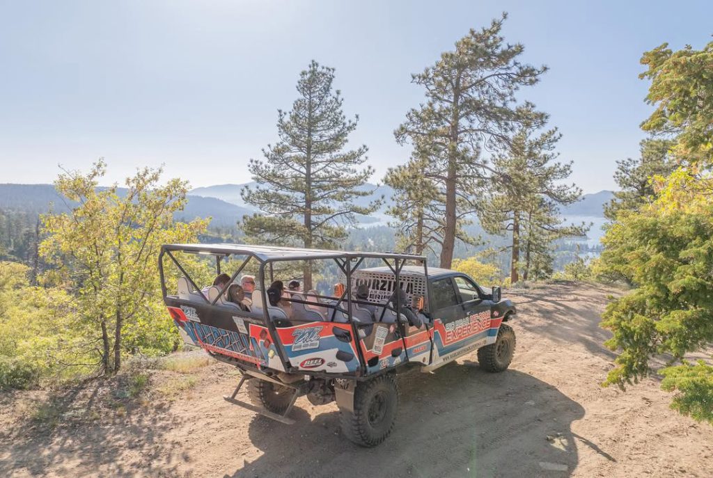 A jeep drives down a dirt road in Big Bear, with several people riding in the back, enjoying the outdoor scenery.