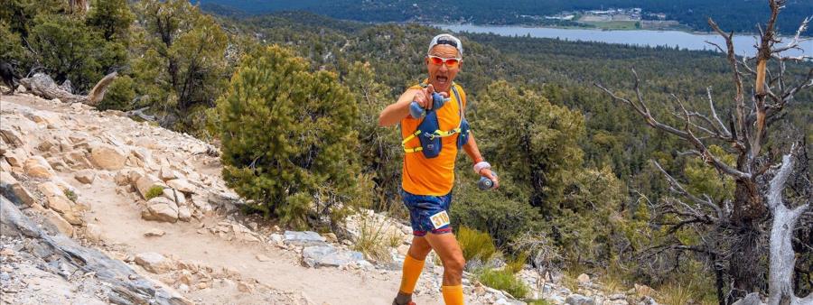 Trail runner wearing orange gear running on a rocky mountain path with forested hills and a lake visible in the background under a clear sky.