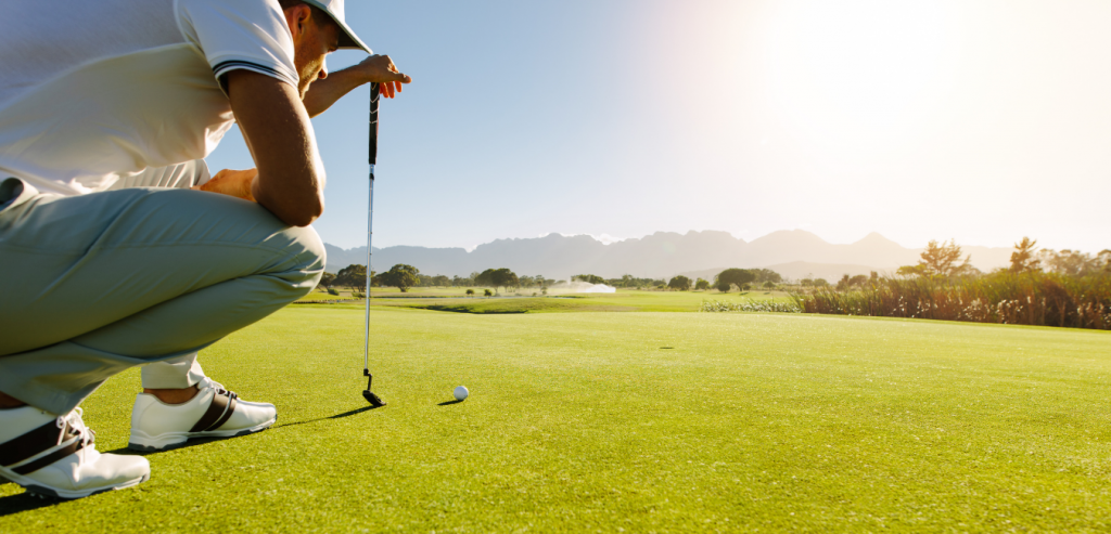 A man in a white polo shirt and light pants crouches on a golf course, lining up a putt on a sunny day with mountains in the distance.
