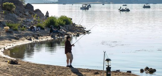 A person stands on the shore, fishing in Big Bear Lake with other boats visible in the distance.