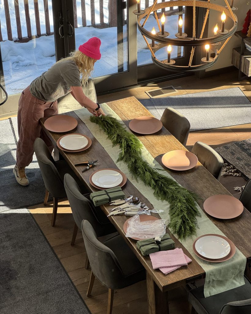 A woman sets a festive Christmas table in a cozy Big Bear production set, surrounded by holiday decorations.