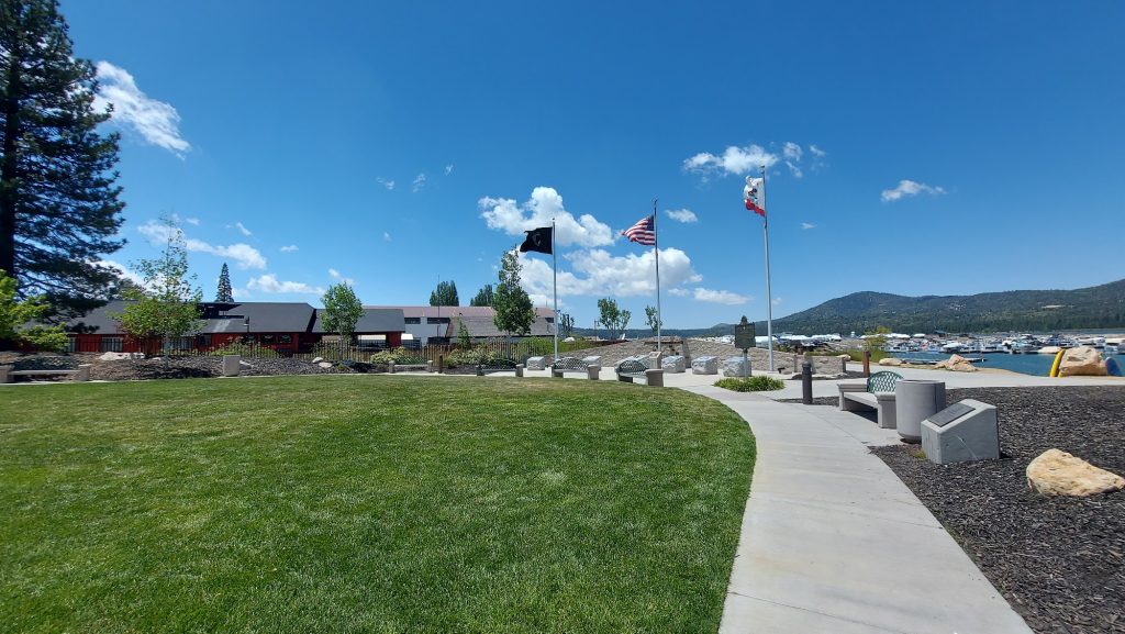Scenic view of Veterans Park in Big Bear Lake featuring manicured lawns, memorial benches, flagpoles, and the marina with mountain views in the background.