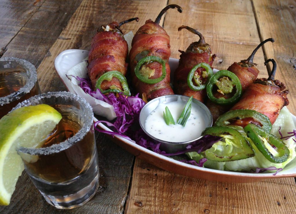 A platter of bacon-wrapped stuffed peppers with sliced jalapeños and a dipping sauce, served with two drinks in salt-rimmed glasses.