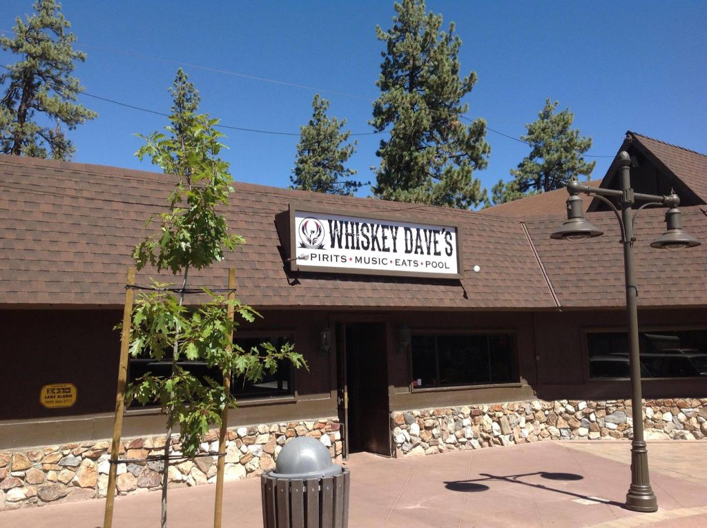 The exterior of Whiskey Dave's at Big Bear, with a brown shingled roof and stone facade, is visible under a clear blue sky.