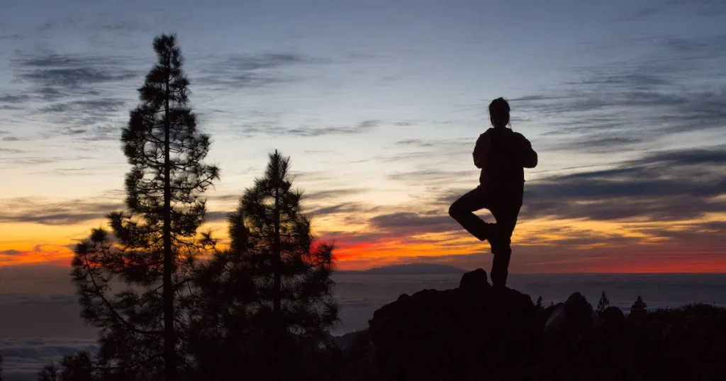 A silhouetted person is doing a tree pose on a mountain at sunset, with a vibrant sky and pine trees in the background at Big Bear